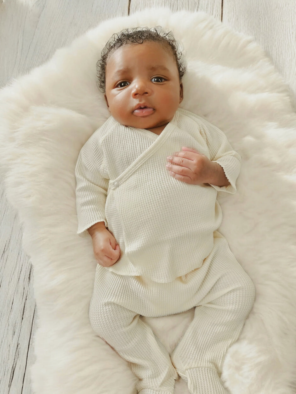 Baby lying on a fluffy white blanket, wearing a cream-colored outfit and looking up with a curious expression.
