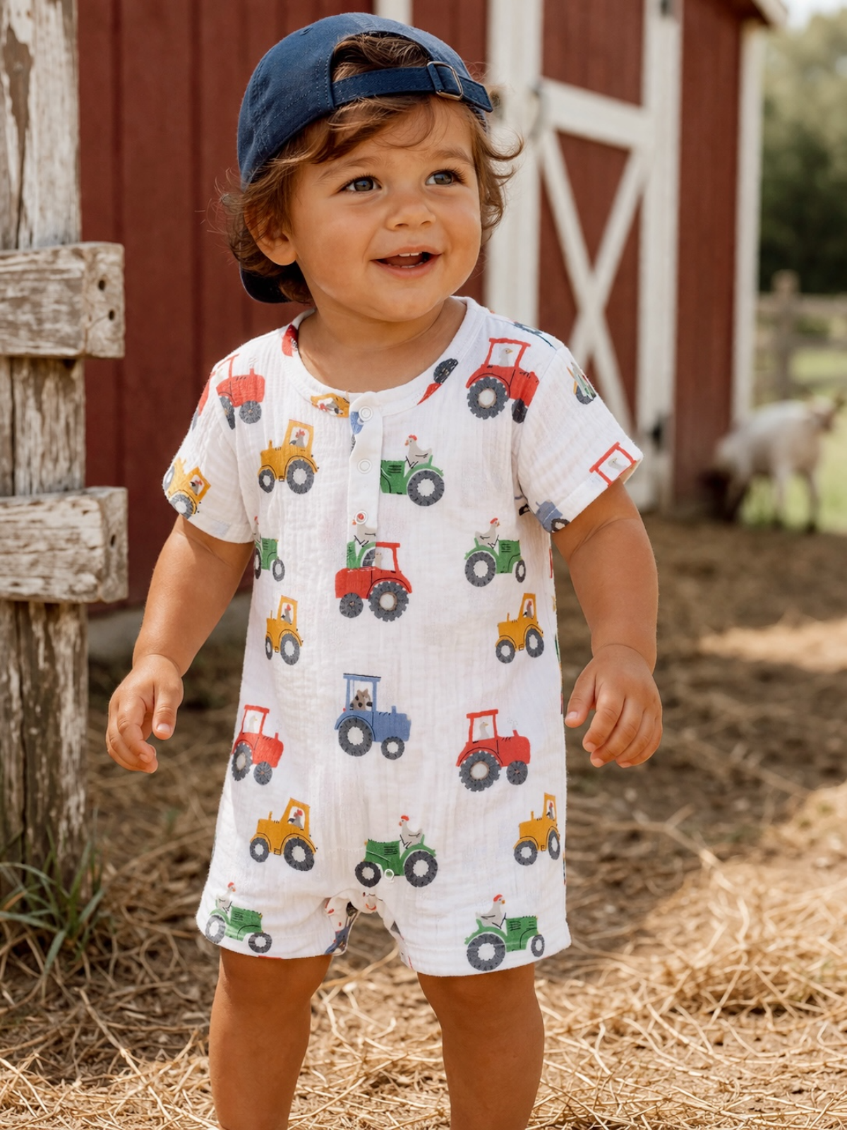 Smiling child in a tractor-patterned outfit stands outside near a barn, wearing a blue cap.