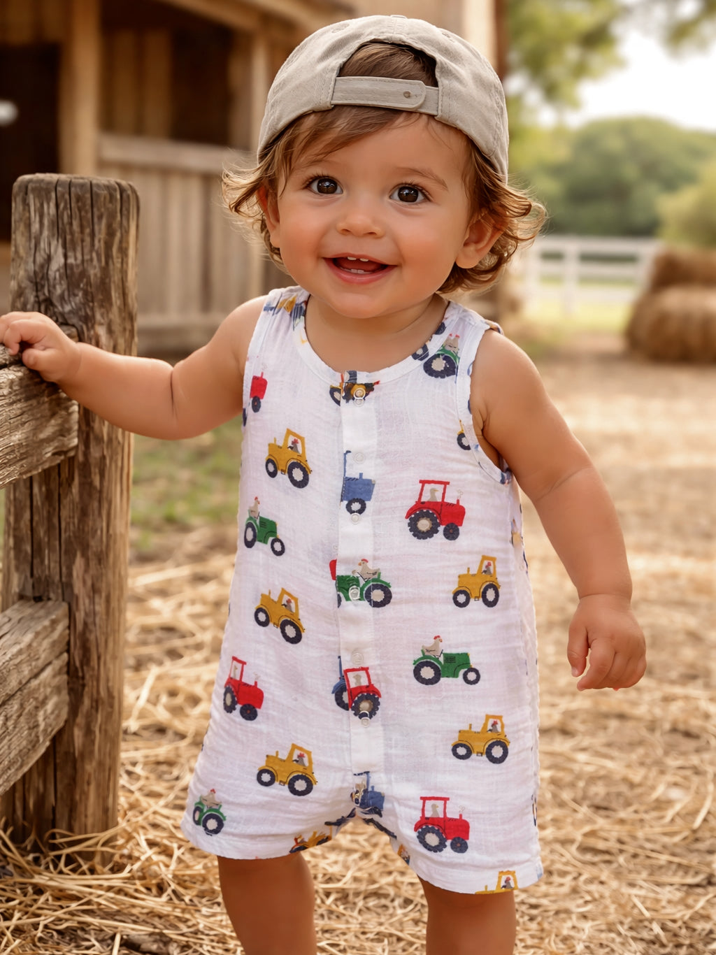 Smiling toddler in a tractor-patterned romper, wearing a cap, standing by a wooden fence on a sunny day.