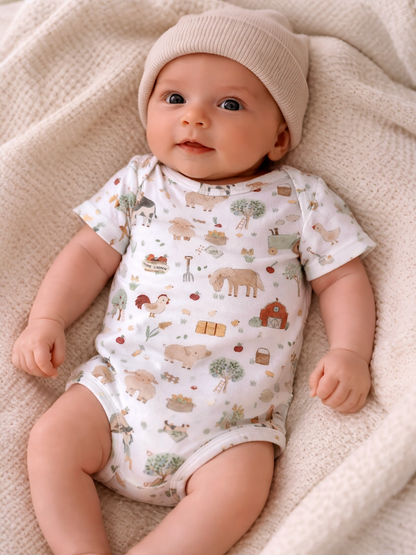 Smiling baby in a light-colored onesie with animal prints, wearing a beige hat, resting on a textured blanket.