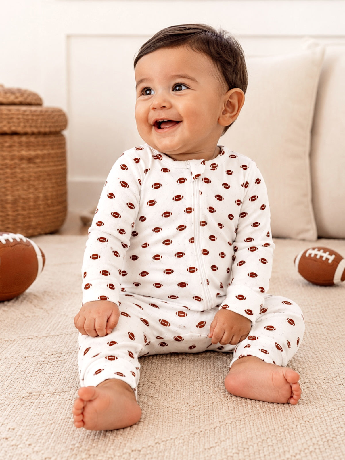 Baby sitting on a rug, smiling, wearing a white outfit with football prints, surrounded by footballs.