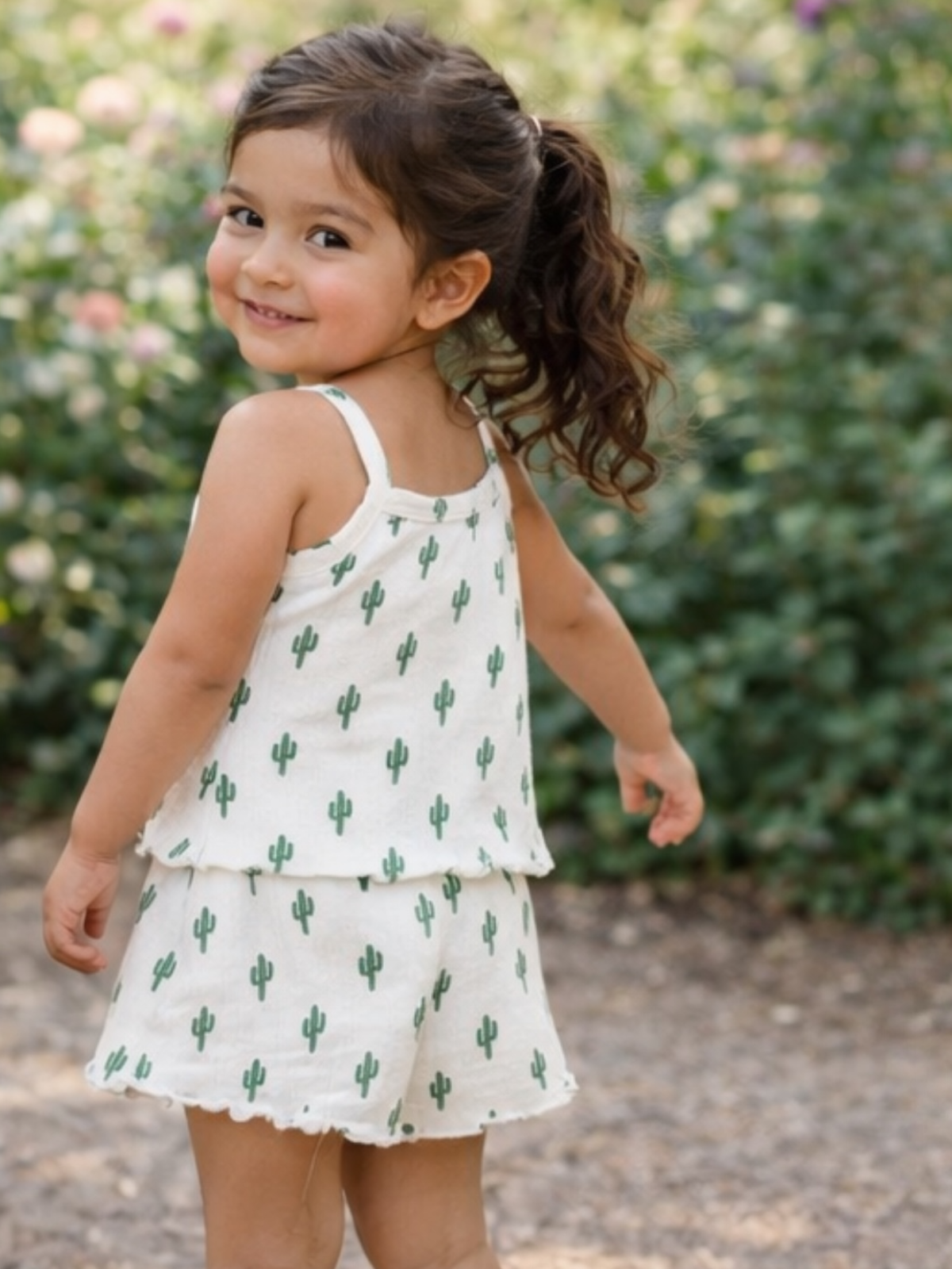 Smiling girl with curly hair in cactus-patterned outfit, standing outdoors among greenery.