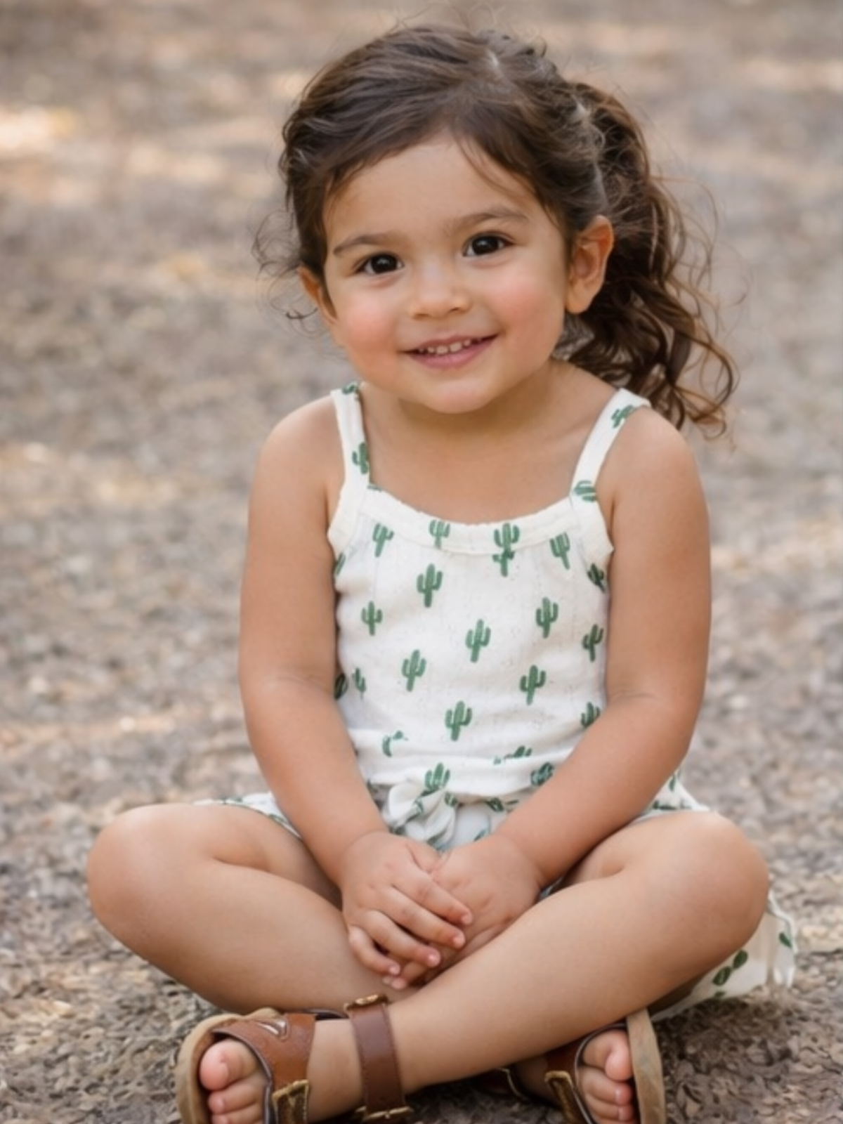 Young girl with long hair, wearing a cactus-patterned dress, sitting and smiling on the ground.