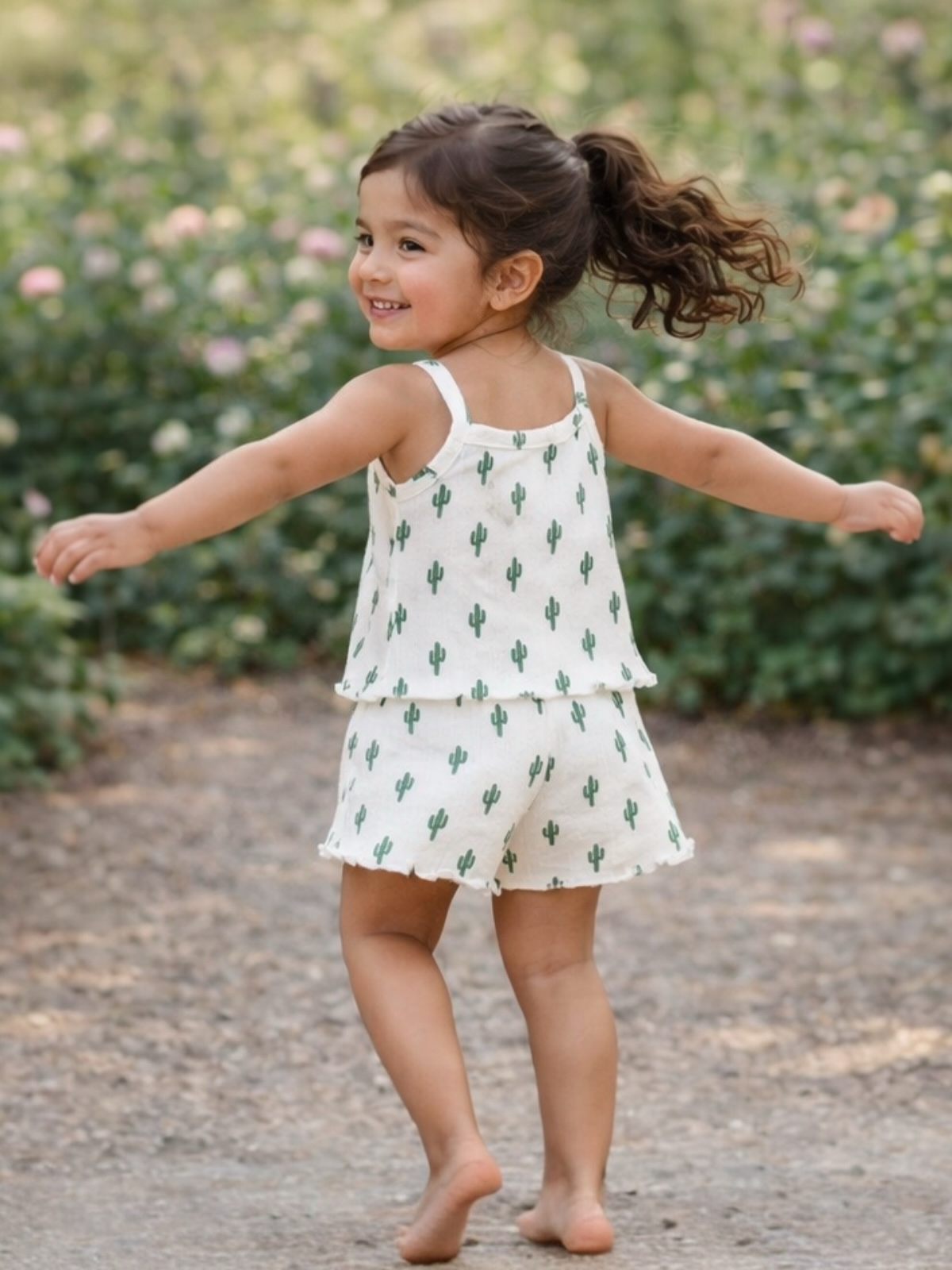 Smiling young girl in cactus-patterned outfit, twirling on a gravel path surrounded by greenery and flowers.