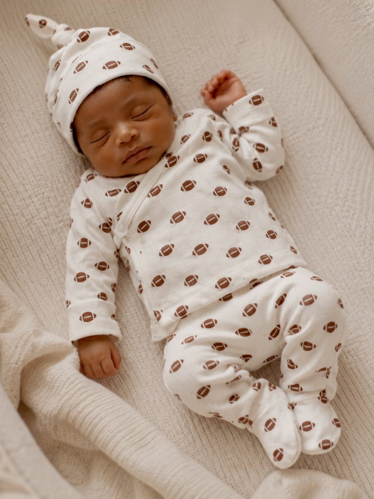Sleeping baby in football-patterned pajamas and hat on a cozy blanket.