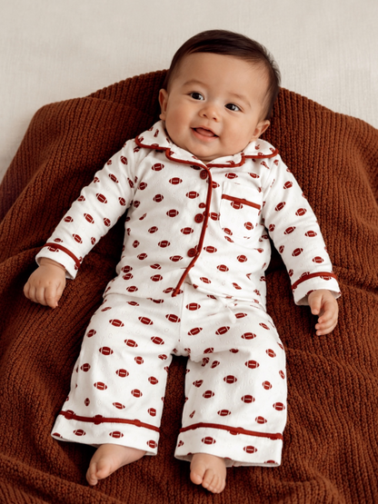 Smiling baby in football-patterned pajamas, sitting on a brown blanket.