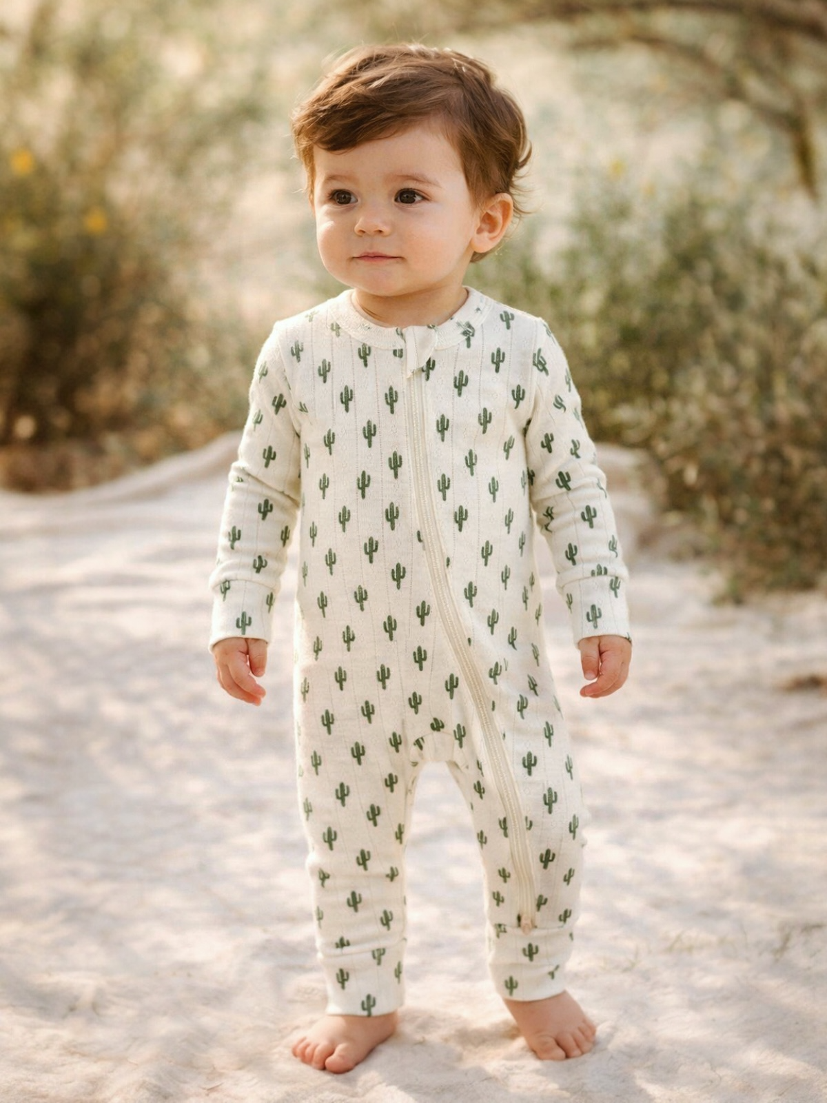 Toddler wearing a cactus-patterned onesie, standing on soft ground, surrounded by greenery.