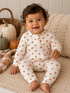 Smiling baby in pumpkin-print pajamas sitting on a cozy blanket with decorative pumpkins in a warm, autumn setting.