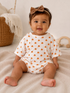 Smiling baby wearing a pumpkin-printed outfit and bow, sitting on a blanket, with a cozy background.