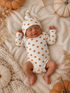 Smiling baby in pumpkin-patterned outfit, surrounded by soft textures and decorative pumpkins.