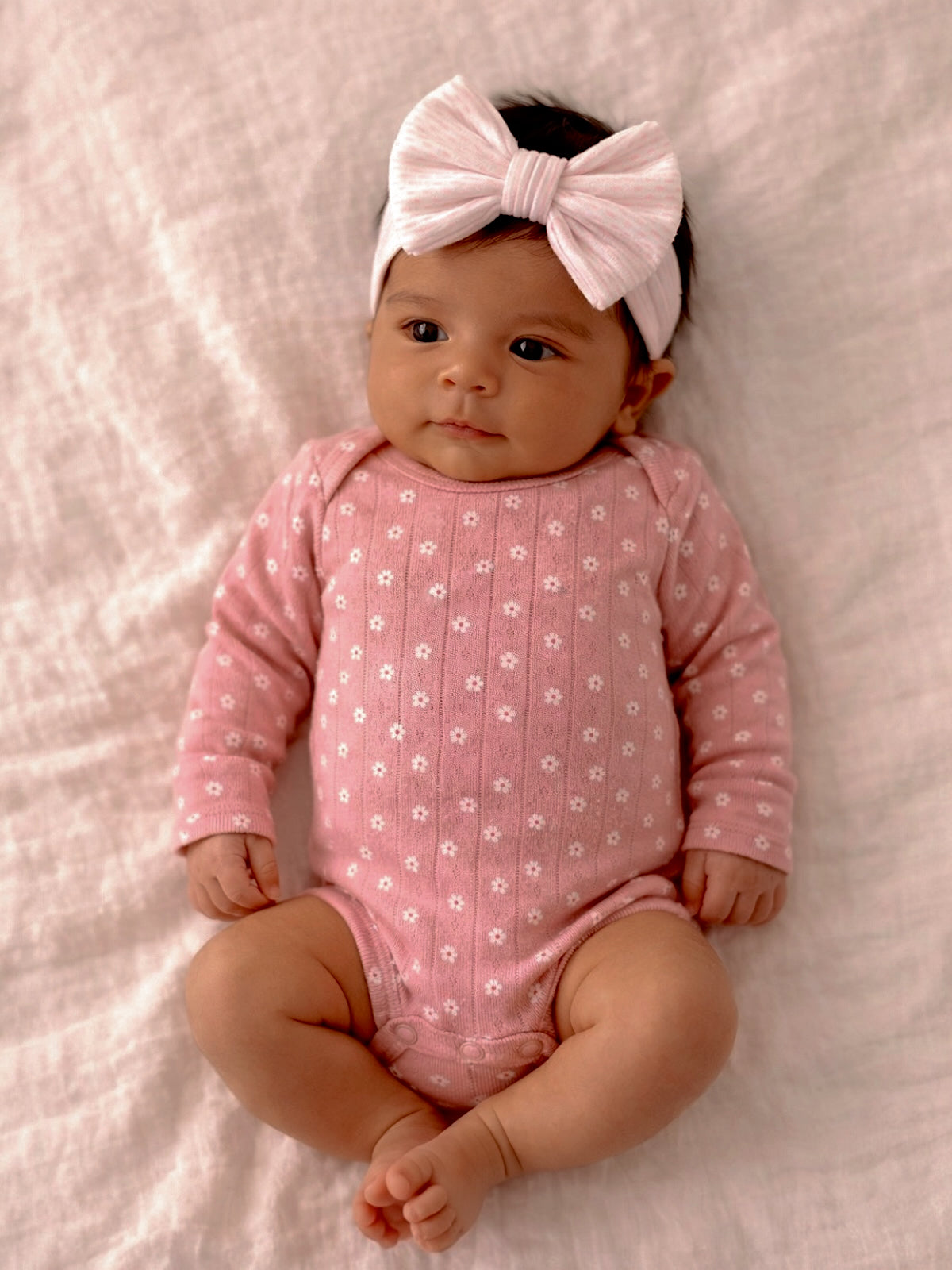 Smiling baby girl in a pink floral onesie and large white bow, lying on a soft white blanket.
