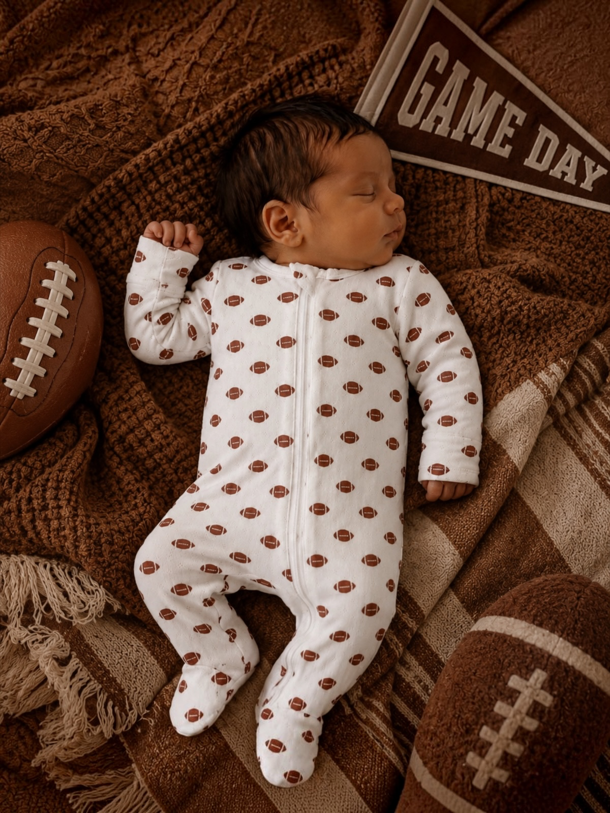 Sleeping baby in a football-patterned onesie, surrounded by footballs and a "Game Day" pennant on a cozy blanket.