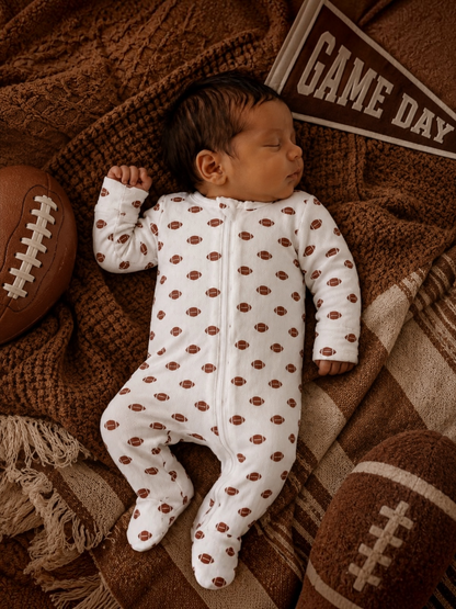 Sleeping baby in a football-patterned onesie, surrounded by footballs and a "Game Day" pennant on a cozy blanket.