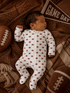 Sleeping baby in a football-patterned onesie, surrounded by footballs and a "Game Day" pennant on a cozy blanket.