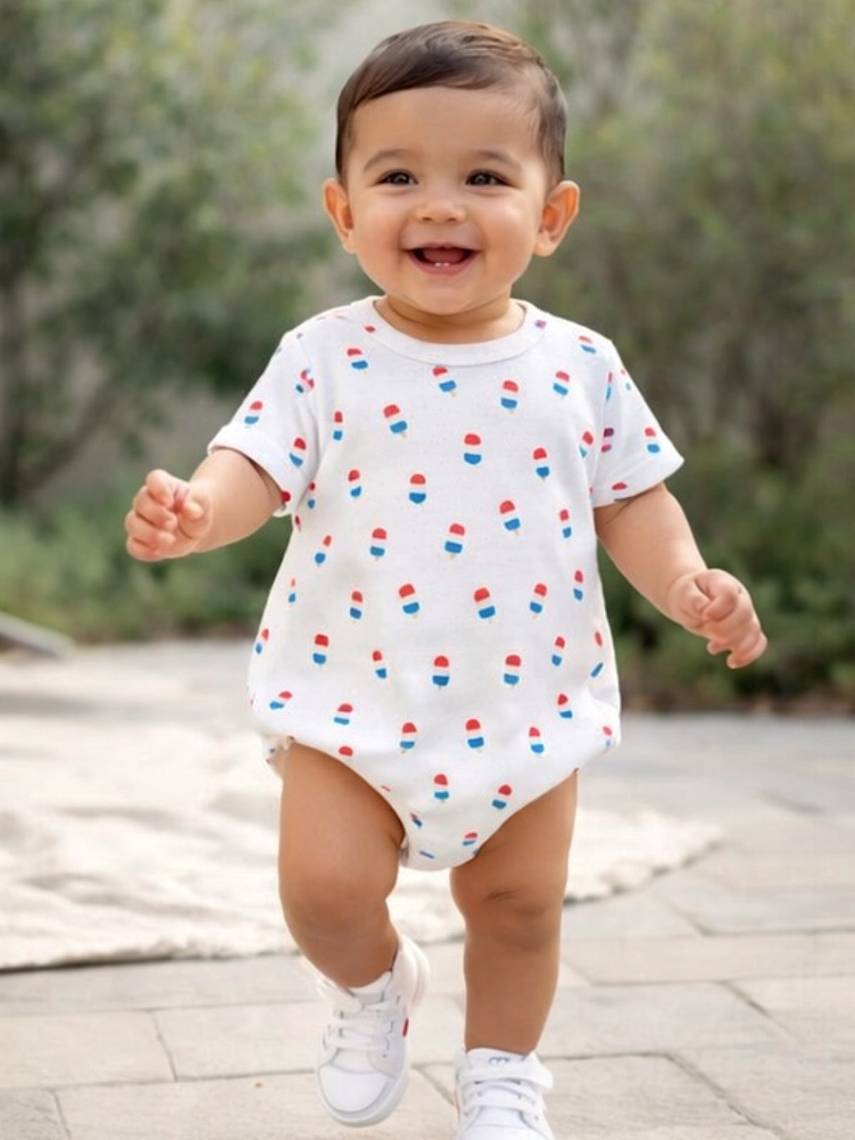 Smiling baby in a patterned onesie walks on a paved outdoor path, wearing white sneakers.