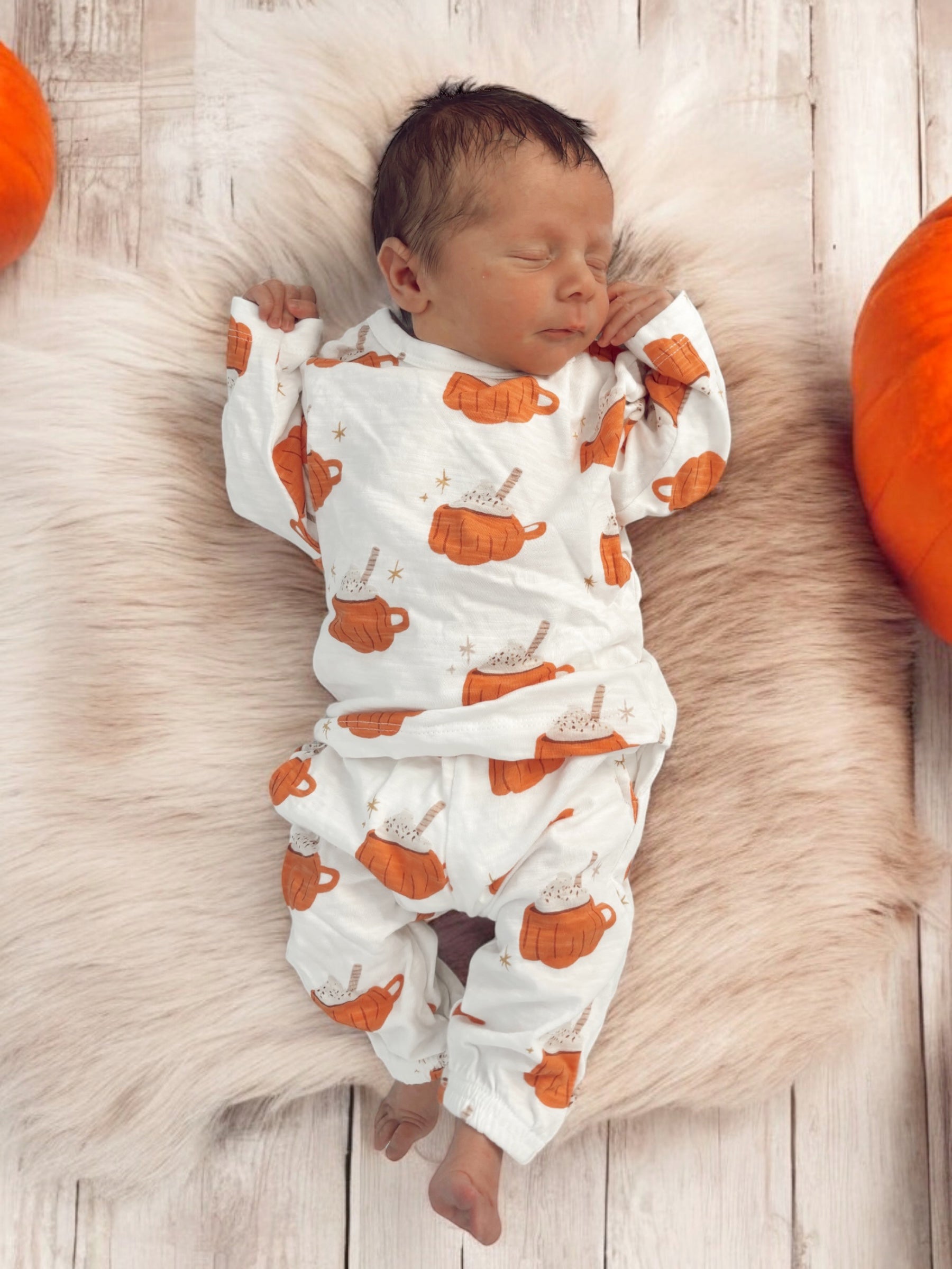 Baby sleeping on a fluffy surface, wearing pumpkin-themed pajamas, surrounded by pumpkins.