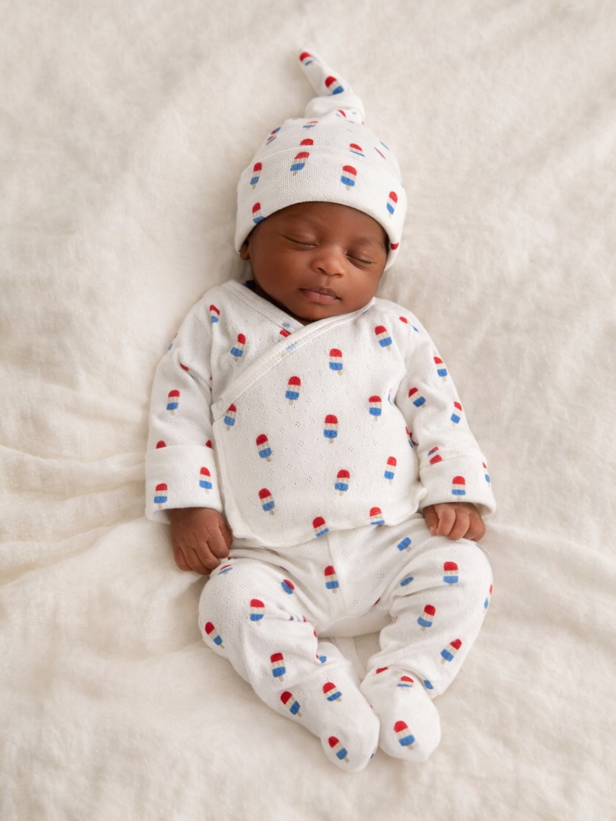 Sleeping baby in white outfit with colorful popsicle patterns, resting on a soft, white blanket.