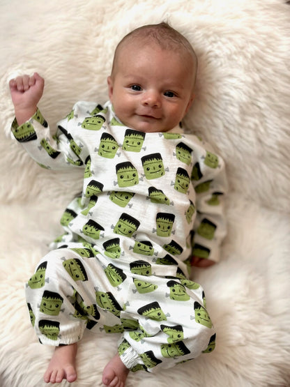 Smiling baby in green Frankenstein-themed pajamas, lying on a fluffy white blanket.