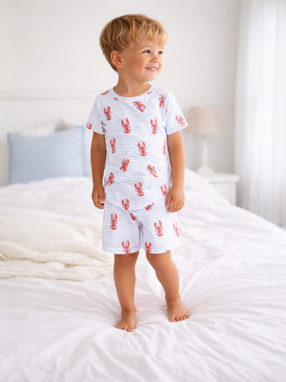 Smiling boy in lobster-patterned pajamas standing on a bed with white bedding and soft natural light.