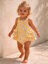 Smiling toddler in a lemon-patterned romper walking on a stone pathway. Natural light and neutral background.