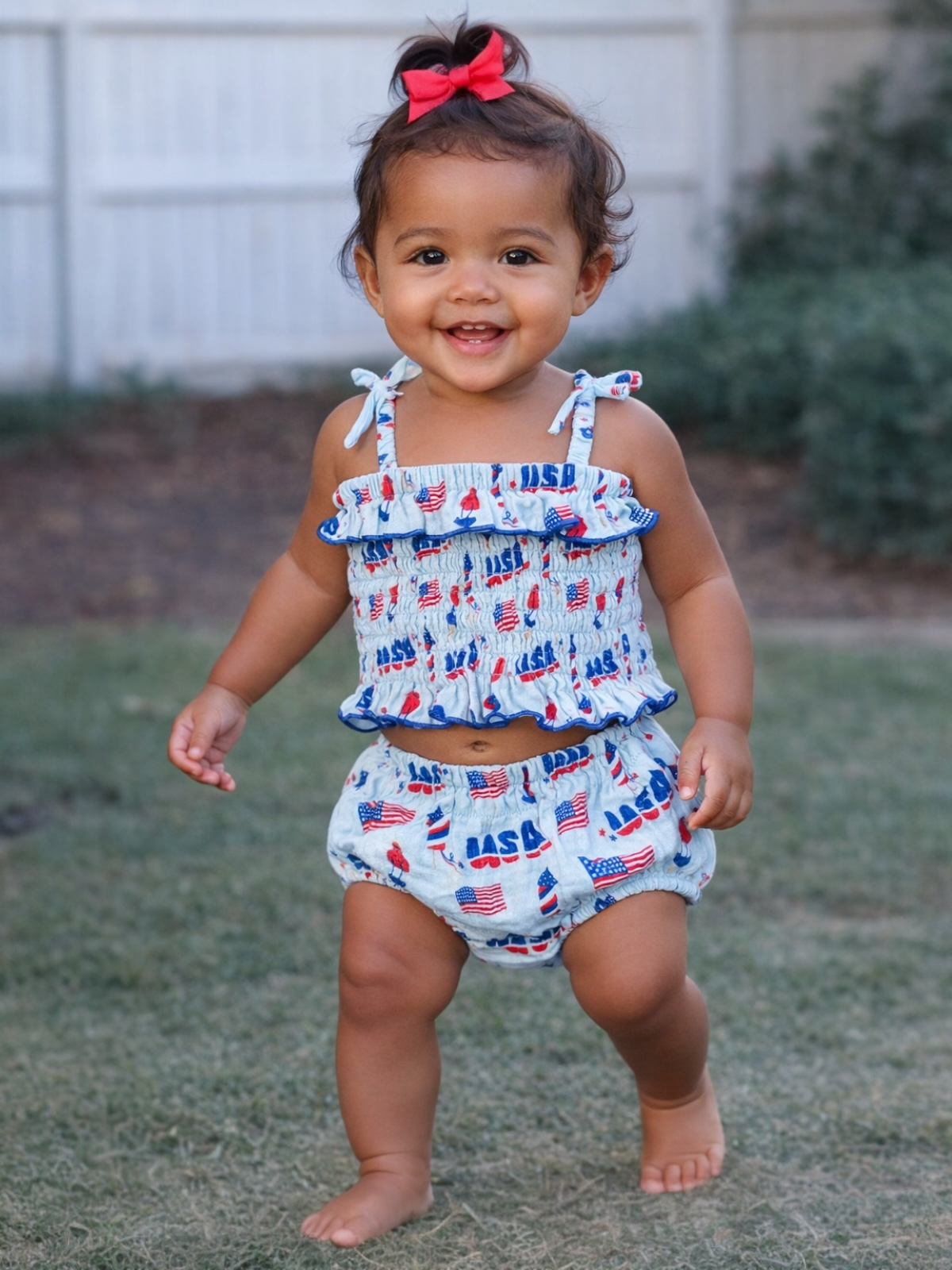 Smiling toddler in a patriotic outfit, walking on grass with a cheerful expression and a red bow in her hair.
