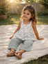 Smiling girl in a floral top and green pants sitting on a blanket in a sunlit outdoor setting.