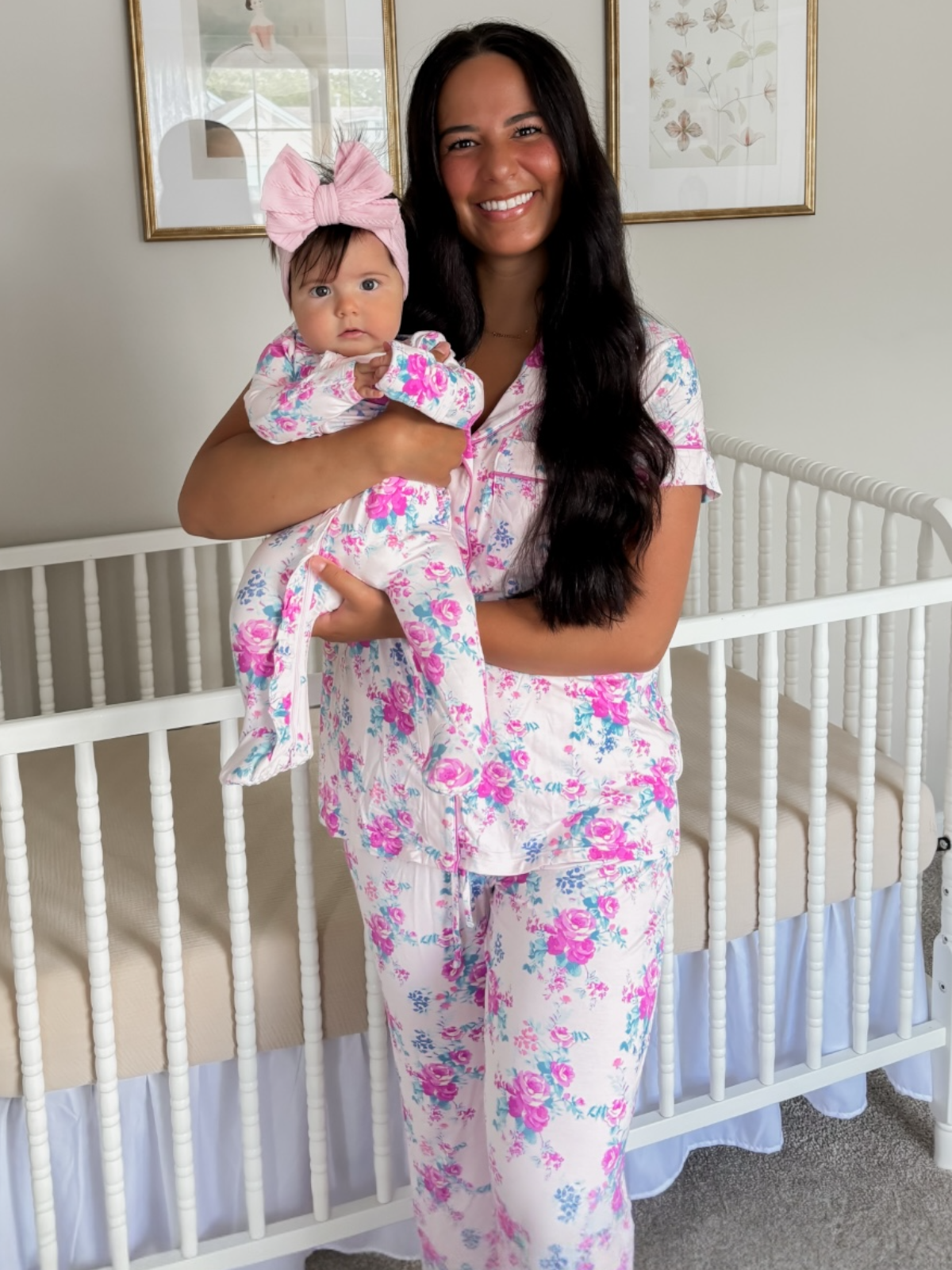 Mother and baby in matching floral pajamas, smiling in a softly lit nursery with a crib in the background.