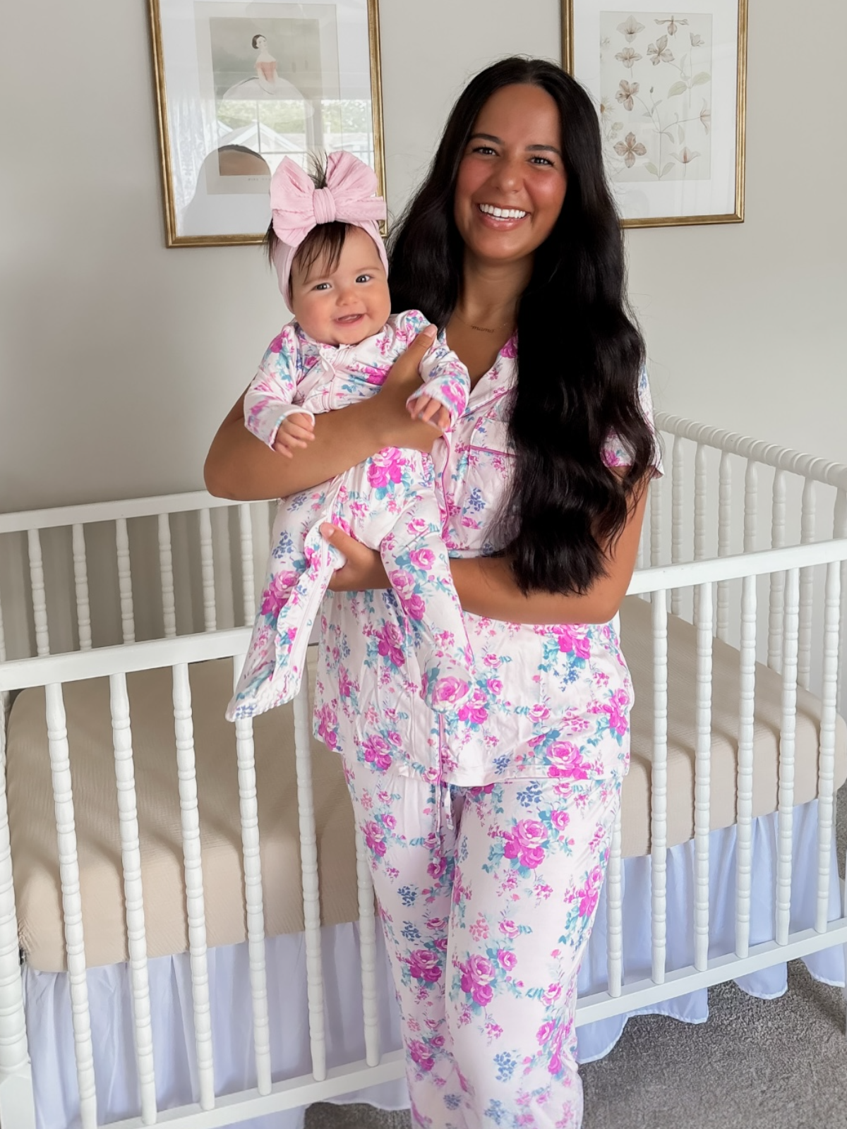 Smiling mother holds a baby in floral pajamas next to a crib in a cozy, well-lit room.