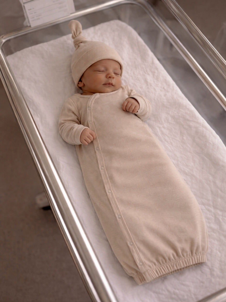 Sleeping newborn in a beige swaddle and knit hat, resting peacefully in a hospital bassinet.