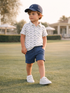 Child walking on grass in a patterned polo shirt, navy shorts, and white shoes, wearing a baseball cap.