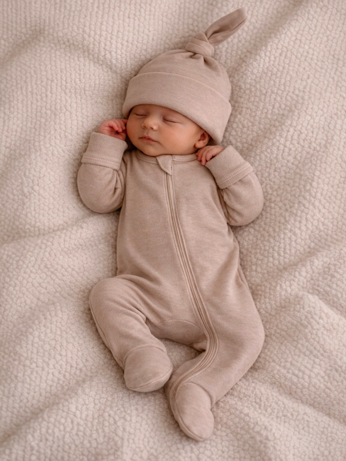 Sleeping baby in a cozy beige onesie and hat, resting on a soft textured blanket.