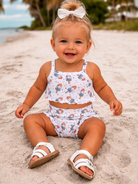Smiling toddler in a popsicle-patterned swimsuit, sitting on sandy beach, with palm trees in the background.