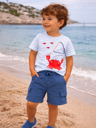 Smiling boy wearing a striped shirt and blue shorts, standing on a beach near water with a crab illustration on his shirt.