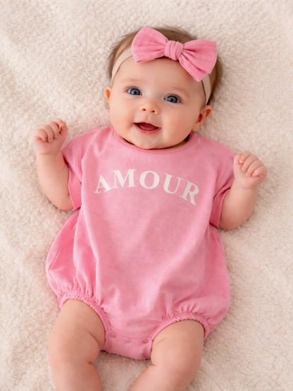 Smiling baby in a pink onesie with "AMOUR" printed, wearing a matching pink bow, on a soft blanket.