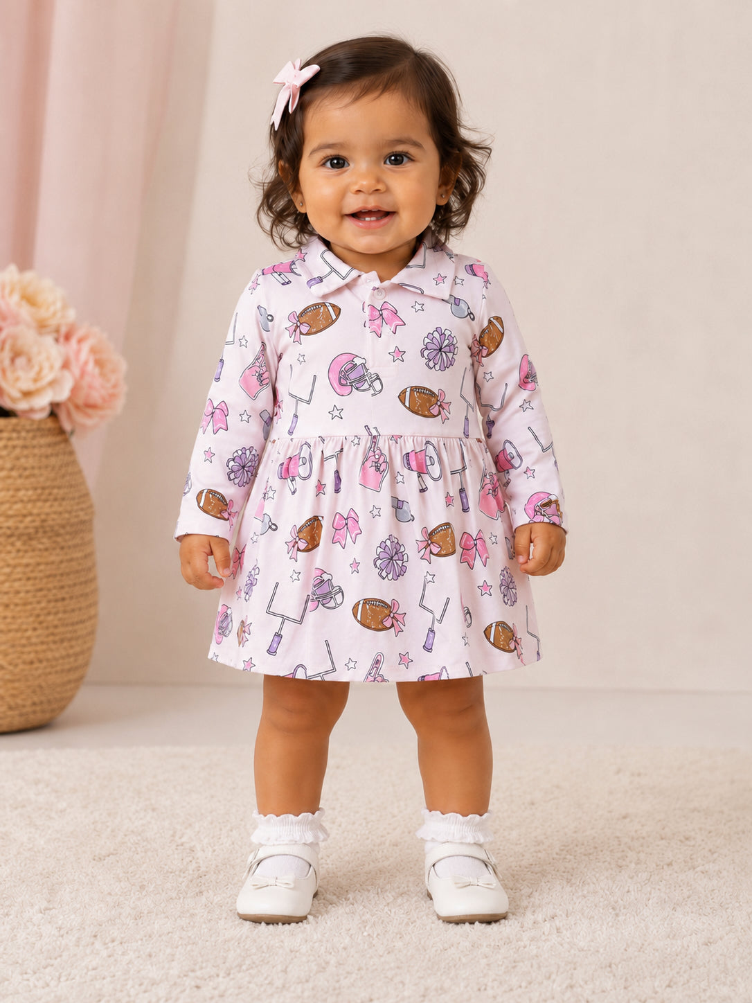 Smiling toddler in a colorful patterned dress with bows and sports themes, standing on a soft rug.