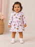 Smiling toddler in a colorful patterned dress with bows and sports themes, standing on a soft rug.