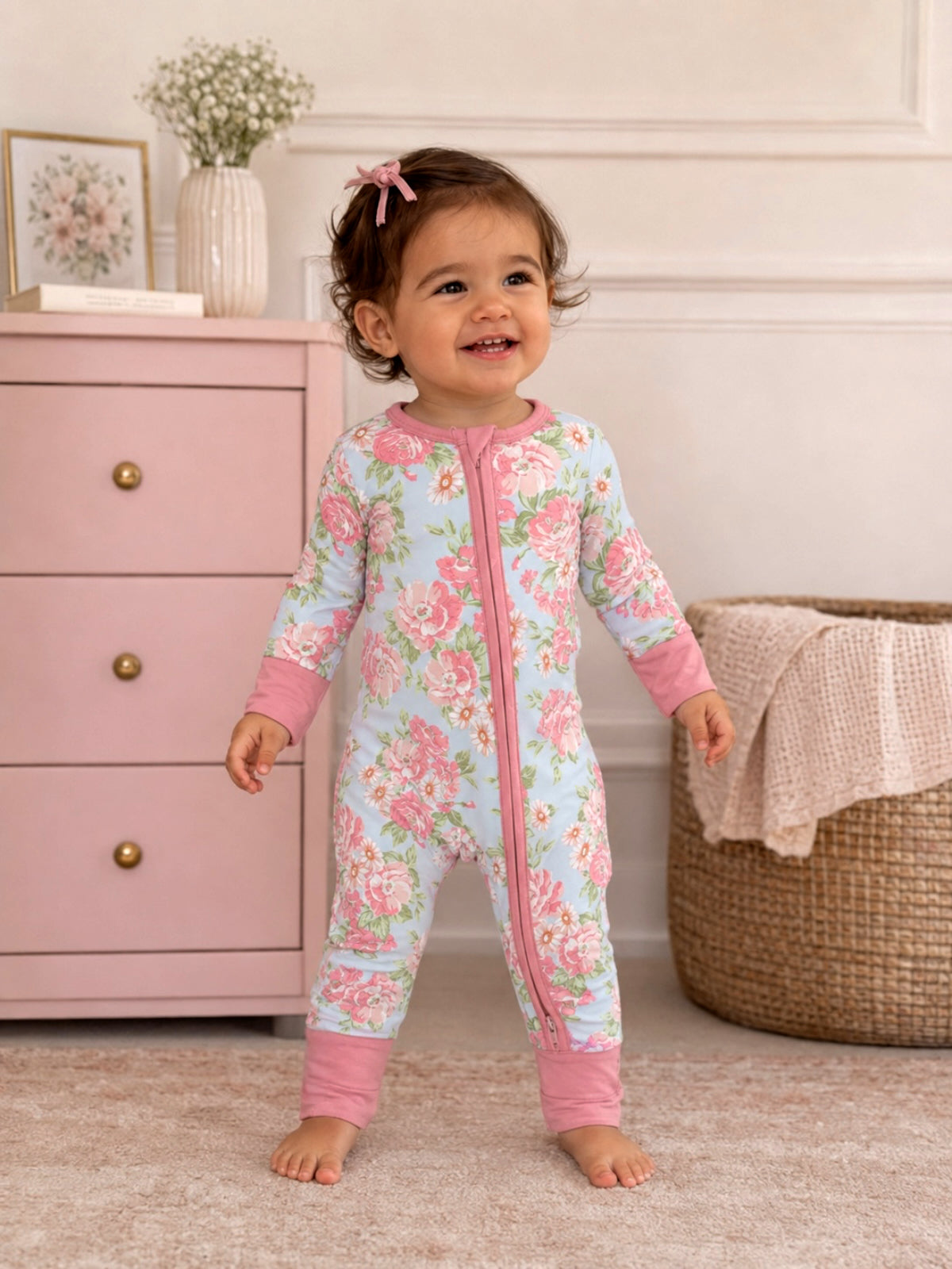 Smiling toddler in a floral onesie stands in a cozy room with pink dresser and braided basket.