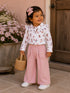 Smiling toddler in a white shirt with colorful boots and pink pants, holding a woven basket near flowers.
