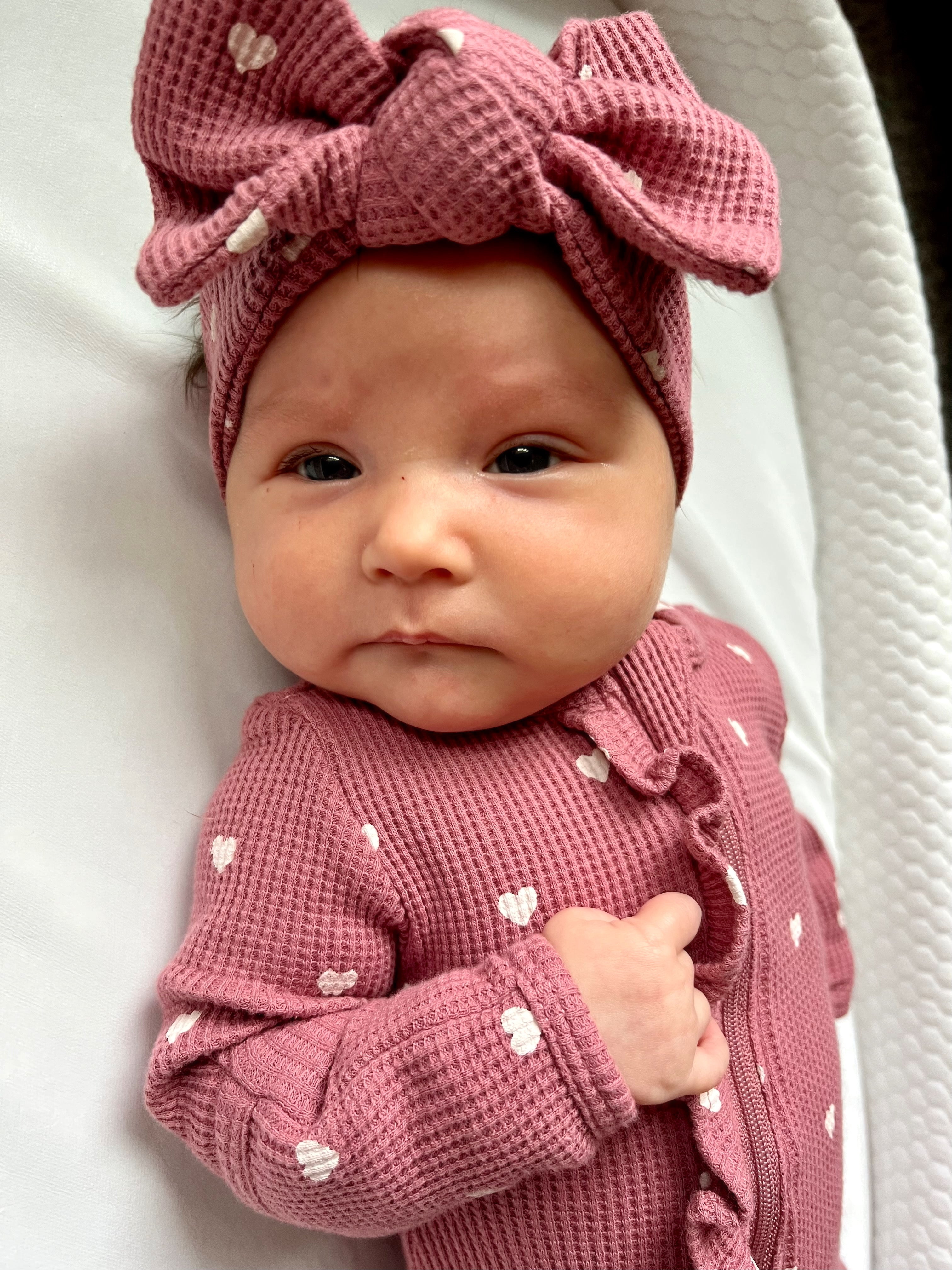 Baby with a pink bow headband and heart-patterned outfit, laying on a white surface, looking at the camera.