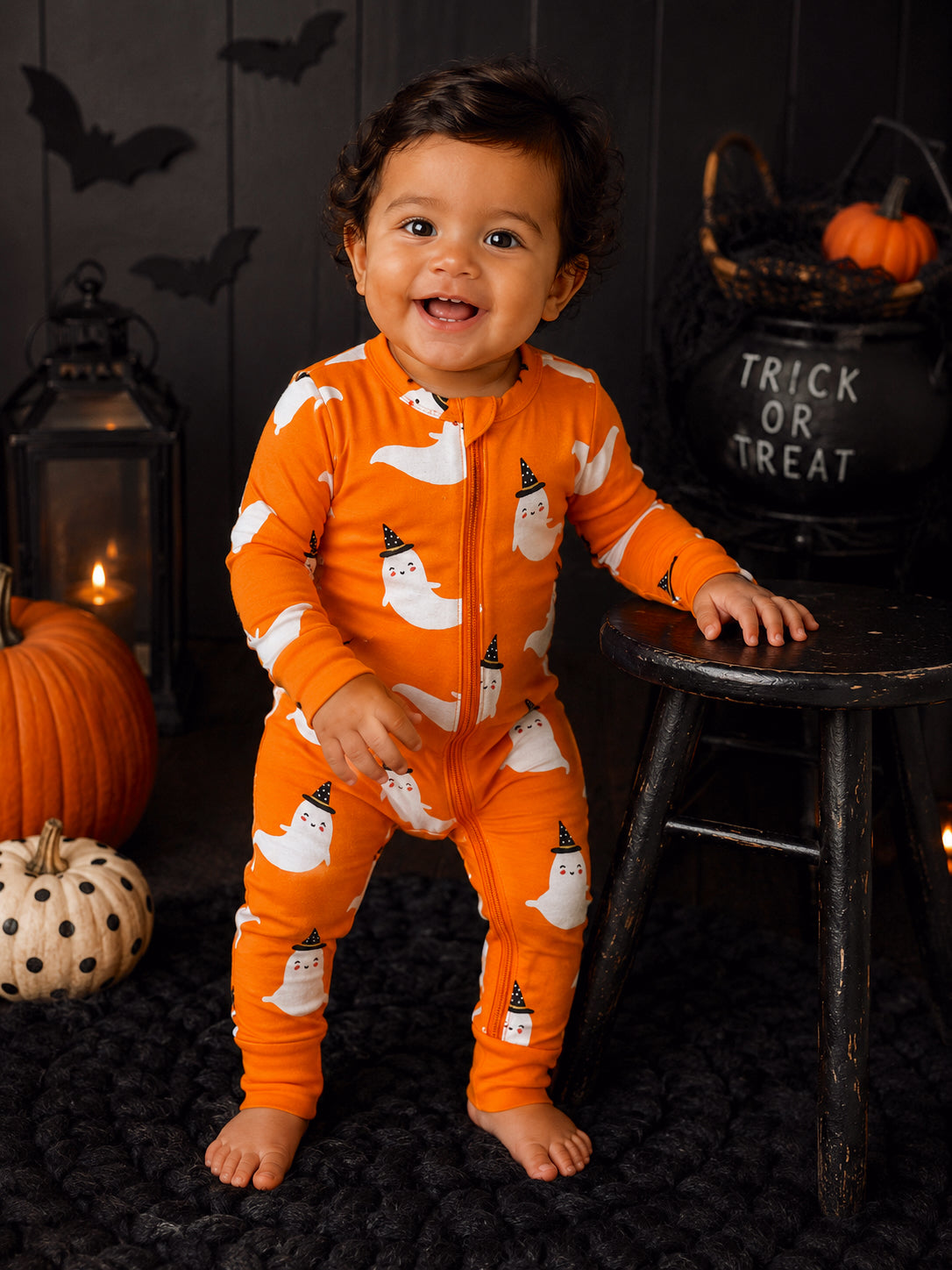 Smiling toddler in orange ghost pajamas, surrounded by pumpkins and Halloween decor, standing next to a wooden stool.