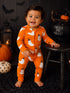 Smiling toddler in orange ghost pajamas, surrounded by pumpkins and Halloween decor, standing next to a wooden stool.