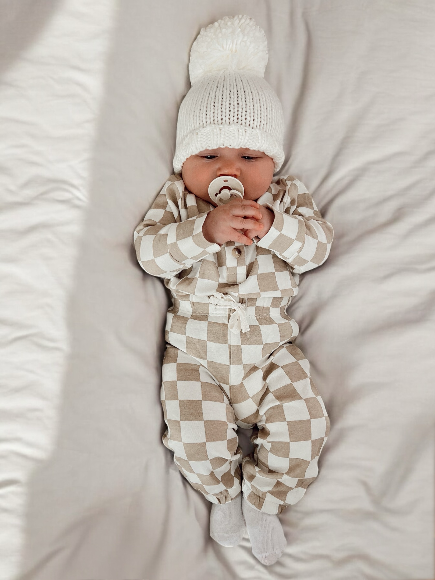 Baby lying on a blanket, wearing a white pom-pom hat and beige checkered outfit, holding a pacifier.