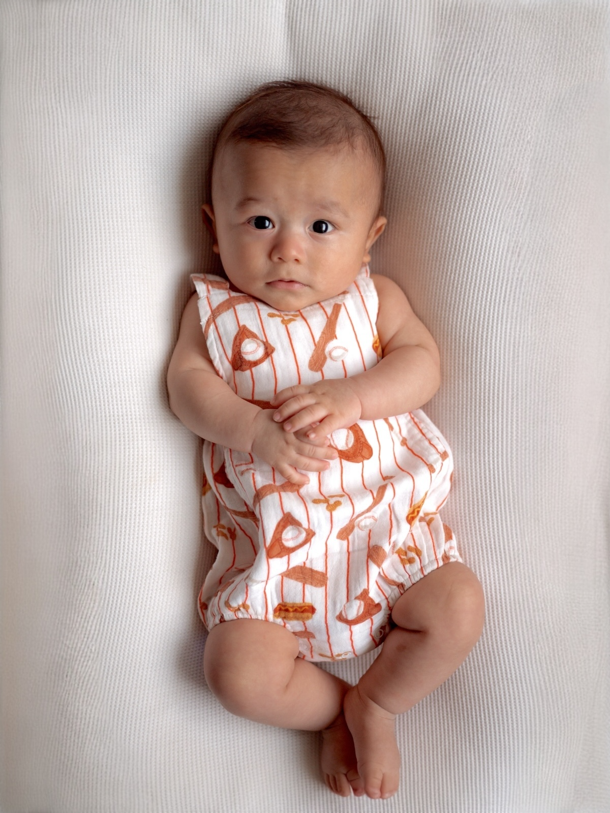Baby in an orange and white outfit lying on a textured surface, looking directly at the camera with hands together.