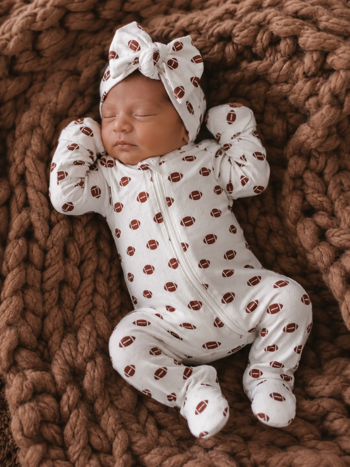 Newborn sleeping in a white football-patterned onesie on a cozy brown blanket.