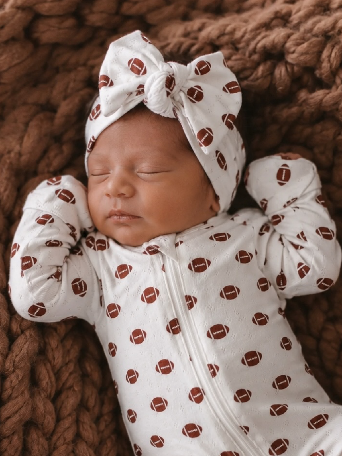 Sleeping newborn in a football-patterned outfit and headband, nestled on a cozy brown blanket.