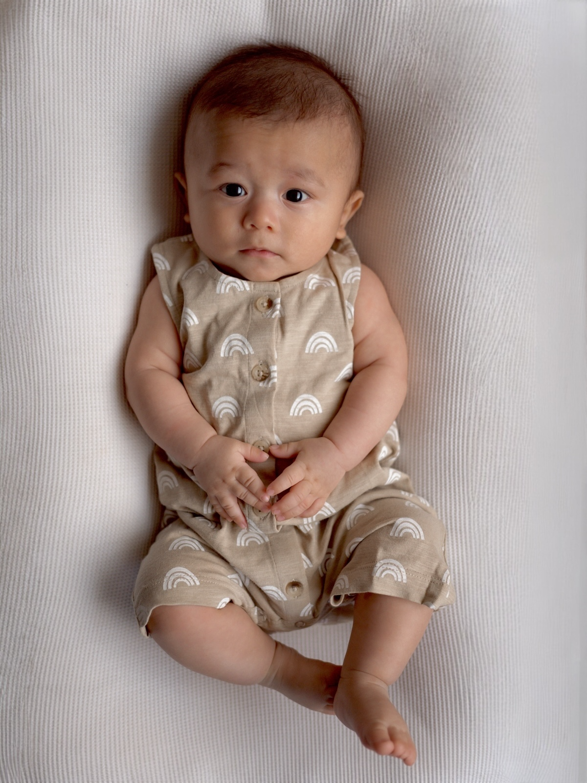 A baby with short hair, wearing a beige outfit with rainbow patterns, sitting on a textured white surface.