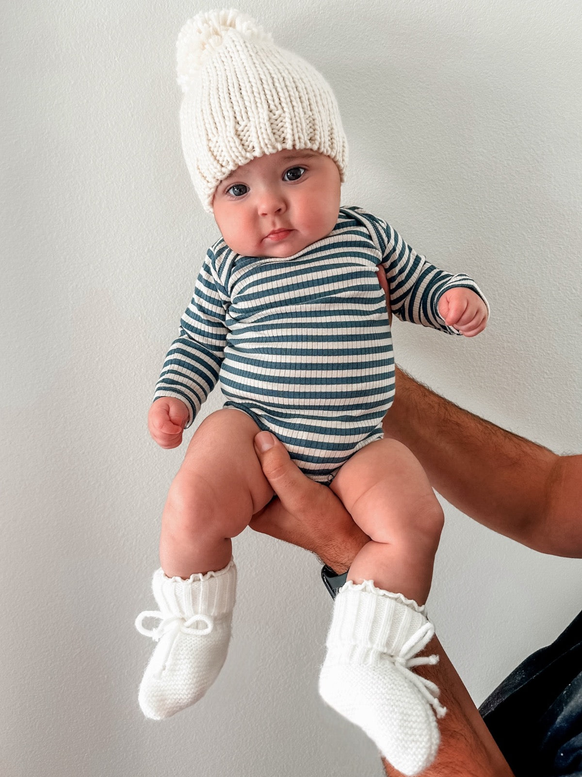 Baby in a white knitted hat and striped outfit, being held by a person against a light background.