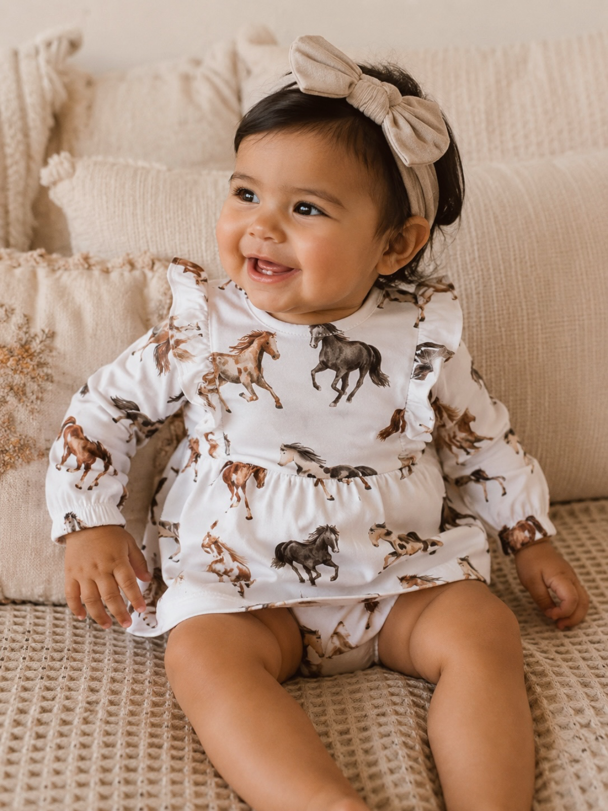Smiling toddler in horse-patterned dress and bow, sitting on a cozy couch with textured pillows.