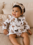 Smiling toddler in horse-patterned dress and bow, sitting on a cozy couch with textured pillows.