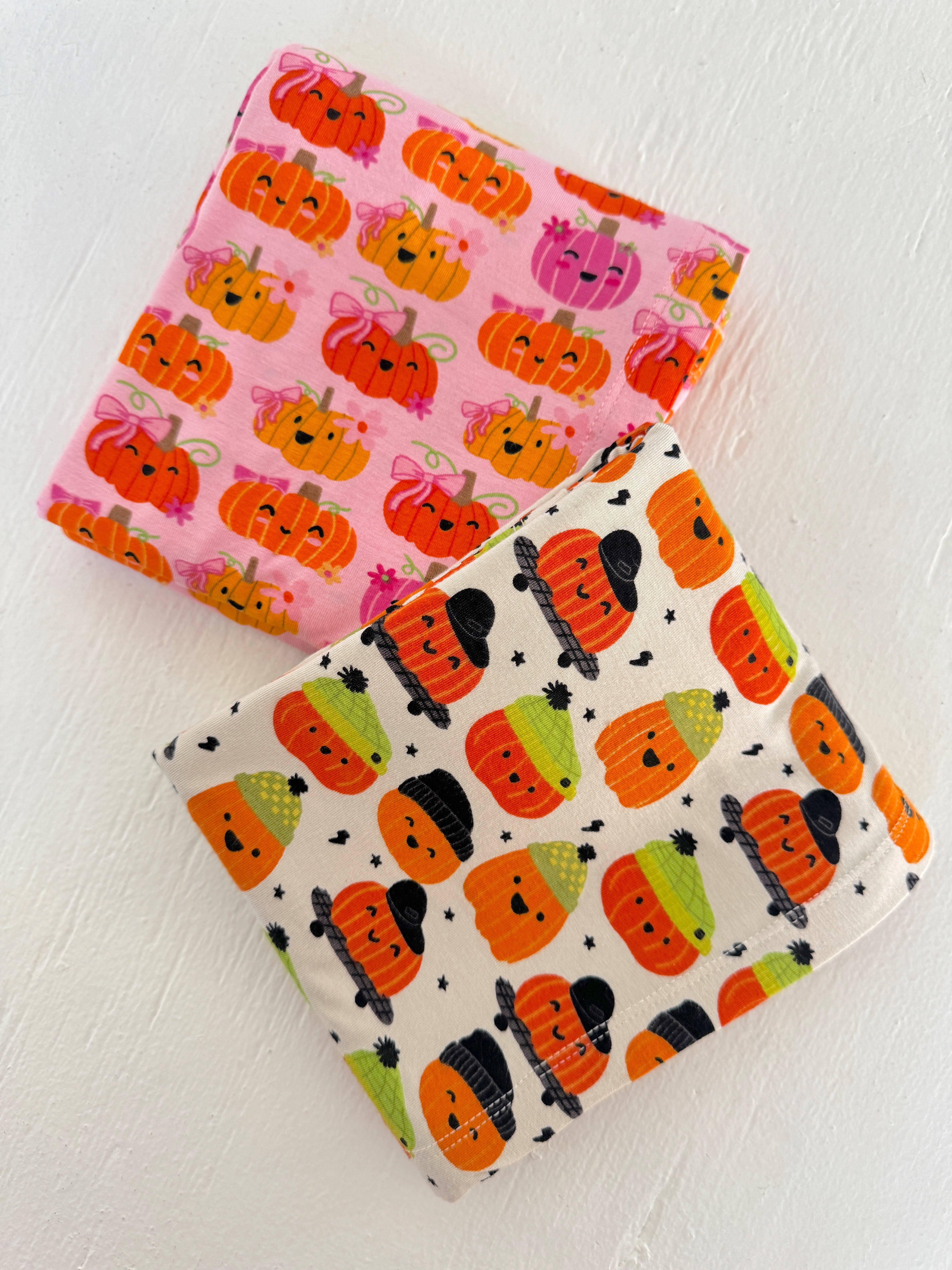 Two folded fabrics with colorful, cartoonish pumpkin patterns in pink and cream colors.